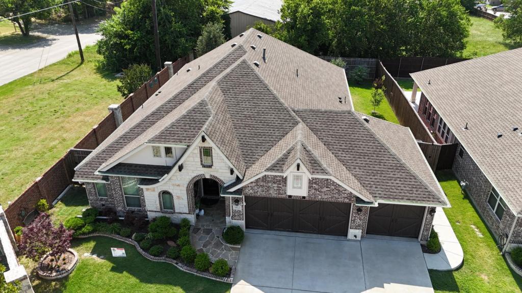 Aerial view of a large, modern home with a brown shingle roof, well-maintained landscaping, and a spacious driveway.